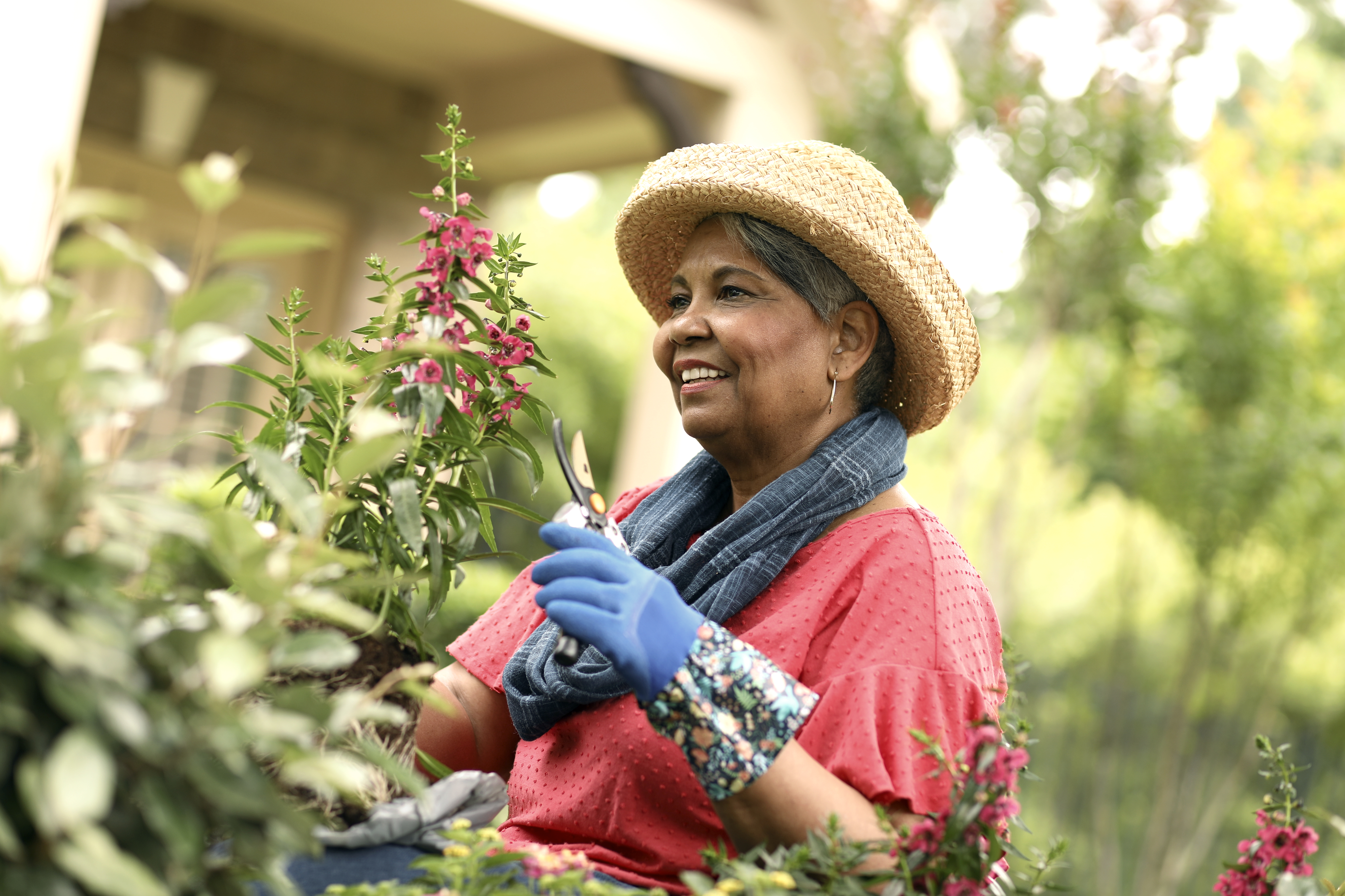 An older woman smiles as she trims flowers in her garden outside.